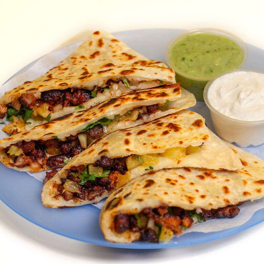 a delicious quesadilla on a blue plate with small containers of sour cream and guac on the side