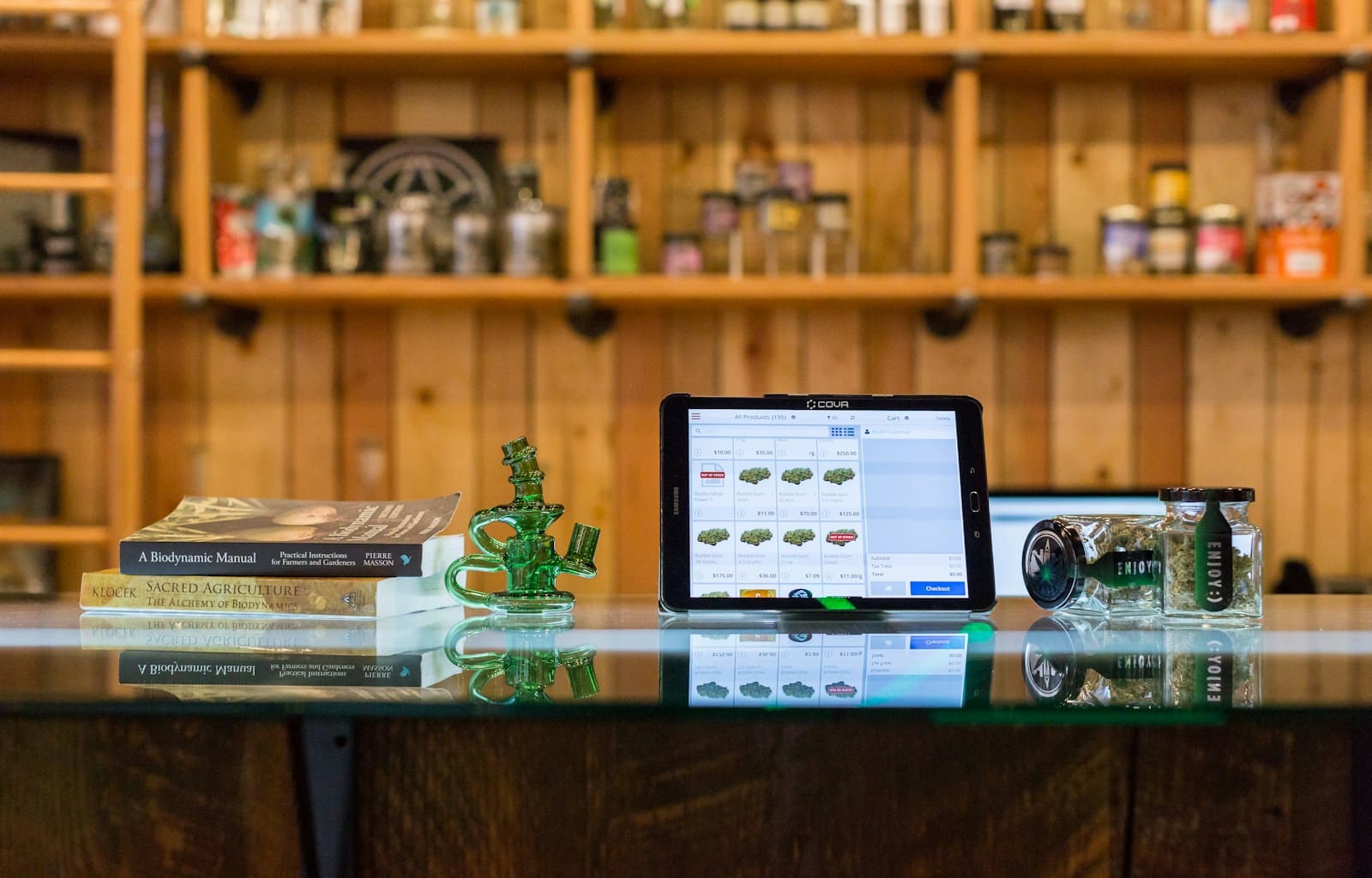 a tablet computer sitting on top of a glass table in a marijuana shop. a green glass bong sits nearby.