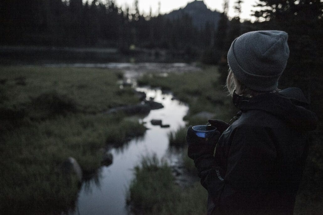 woman standing beside water and coniferous trees. Pinene is a naturally occurring chemical in both pine trees and cannabis.