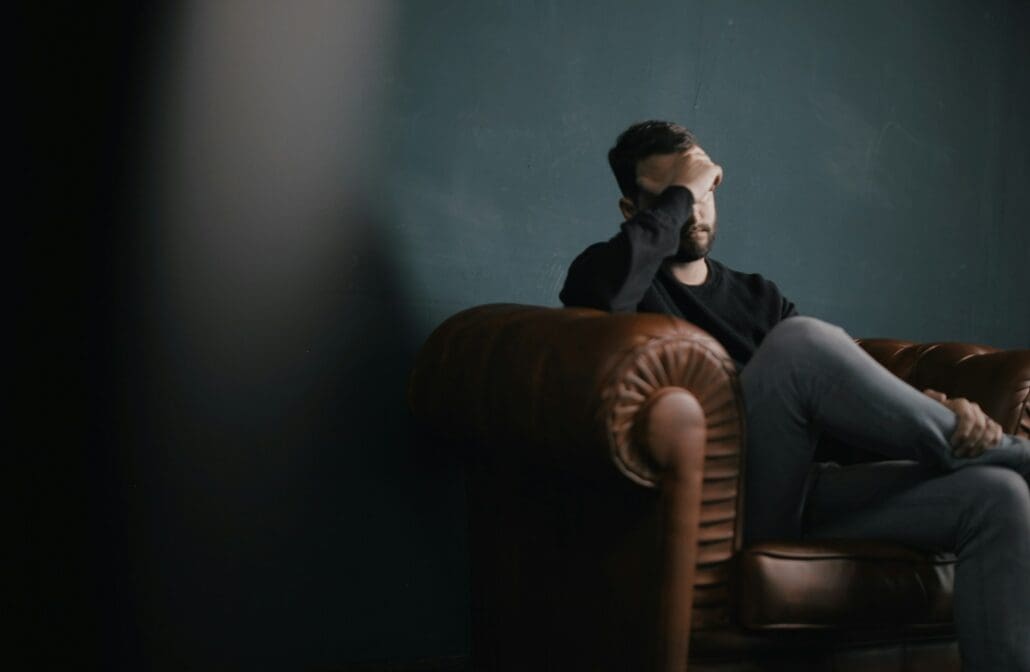 a white man with dark hair holds his head while sitting on a sofa in semi-darkness. 