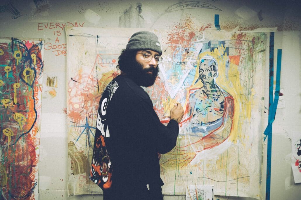 man an artist in black and white sweater standing beside wall with graffiti influenced art. He wears a gray beanie and has a bush black beard and mustache