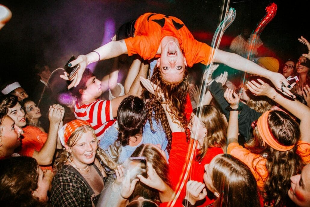a group of costumed people standing around each other at a Halloween party in Tacoma. A woman in a jack o lantern short is crowd surfing.
